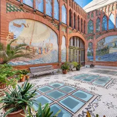 Patio de la Casa Navàs de Reus, con murales cerámicos que narran historias marítimas y paisajes, suelo de mosaico y plantas.