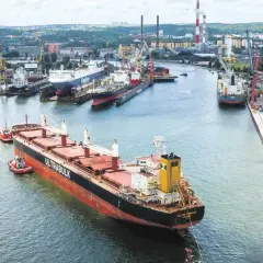 Duży statek towarowy "ULTRABULK" manewruje w porcie z pomocą holowników, w tle stocznie i miasto.