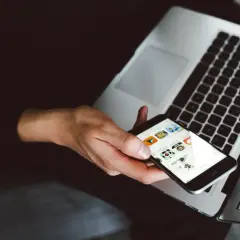 Dłoń trzyma iPhone'a z widocznymi ikonami aplikacji, obok otwartego laptopa. To idealny moment na **backup iPhone'a**.