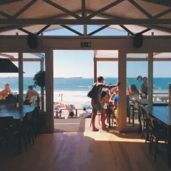 Widok z wnętrza restauracji na plażę i morze. W środku ludzie siedzą przy stołach, na zewnątrz plażowicze odpoczywają. To najlepsza restauracja na Helu.