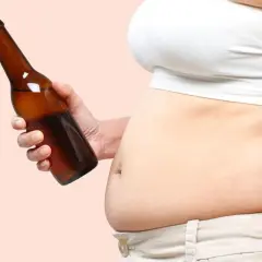 Dłoń trzyma butelkę piwa, obok widoczny brzuch piwny.