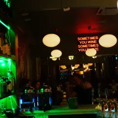 En livlig bar med grönt ljus som lyser upp hyllor med flaskor. En neonskylt med texten "SOMETIMES YOU WINE SOMETIMES YOU POZE" hänger ovanför en bar där gäster sitter. Tapas 28.