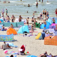 Tłum ludzi na plaży, opalający się i kąpiący w morzu, kolorowe parawany i namioty plażowe.