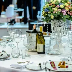 Elegancko nakryty stół w restauracji, gotowy na przyjęcie weselne. Na białym obrusie stoją kieliszki, butelka wina i piękne kwiaty.
