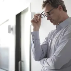 Mann mit Brille, der sich an eine Wand lehnt. Er wirkt nachdenklich, vielleicht kämpft er mit Hitzewallungen.