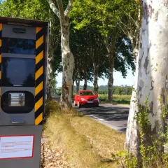 Panneau de **détecteur de radar interdit** sur le bord de la route, avec une voiture rouge passant à proximité.