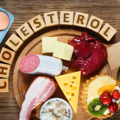 Drewniane klocki z napisem "CHOLESTEROL" otaczają jajka, masło, wątróbkę, kiełbasę, boczek, ser, sardynki i ciastko z owocami.