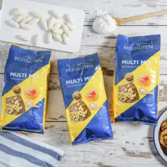 Trzy opakowania mieszanki bezglutenowej Multi Mix, placuszki, mąka i białe paluszki na desce.