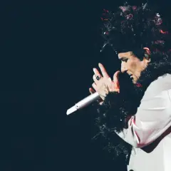 Renato Zero, con un look teatrale e piume nere, canta sul palco, lontano dalle spiagge.