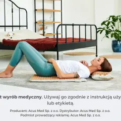 Kobieta leży na macie akupresurowej, która zapewnia masaż uda. W tle łóżko z psem.