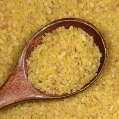 Drewniana łyżka pełna drobnej, żółtej kaszy bulgur na tle rozsypanych ziaren. To zdrowy składnik wielu dań.
