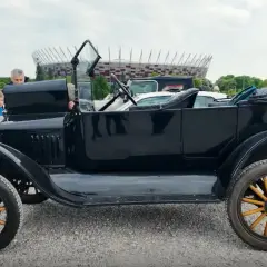 Czarny, zabytkowy Ford T z drewnianymi szprychami kół, obok którego stoją ludzie. W tle widać stadion.