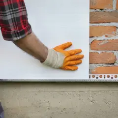 Pose d'isolant blanc sur un mur de briques. La main gantée ajuste la plaque, une étape clé dans la définition de l'isolation.