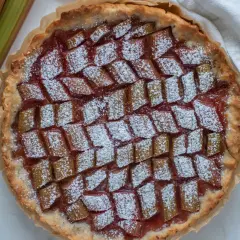Pyszna tarta z rabarbarem, posypana cukrem pudrem, obok świeże łodygi rabarbaru.