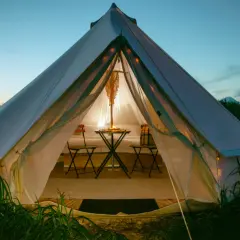 Przytulna glamping jurta oświetlona od środka, z małym stolikiem i krzesłami, otoczona zieloną trawą o zmierzchu.