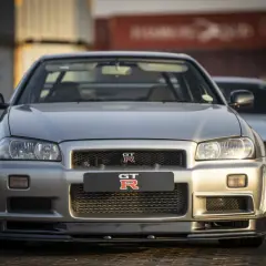 Srebrny Nissan Skyline GT-R R34 z bliska, z innym GT-R w tle. Ikoniczny samochód sportowy.