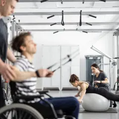 Rehabilitacja metodą Bobath: mężczyzna na wózku ćwiczy z fizjoterapeutą, kobieta na piłce z hantlami pod okiem terapeutki.