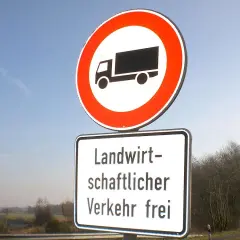 Verkehrsschild: LKW verboten, aber landwirtschaftlicher Verkehr frei. Blaue Himmel und Bäume im Hintergrund.