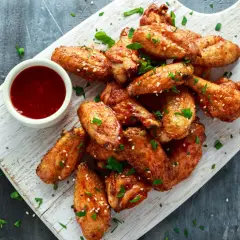 Alitas de pollo al horno jugosas, doradas y espolvoreadas con sésamo y perejil, acompañadas de una salsa roja en un cuenco.