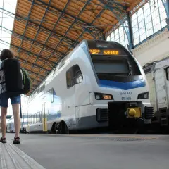 Zwei Reisende mit Rucksäcken warten auf den serbischen Zug nach Szeged unter dem beeindruckenden Dach eines Bahnhofs.