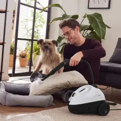 Mann reinigt Kissen mit Dampfreiniger, während Hund zusieht. Eine einfache Methode für sessel reinigen hausmittel.
