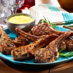 Chuletillas de cordero a la parrilla, jugosas y doradas, servidas en un plato azul con romero fresco y una salsa amarilla.