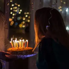 Wnuczka zdmuchuje świeczki na torcie urodzinowym. W tle migoczą światełka, tworząc magiczną atmosferę.