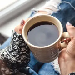 En person med slitna jeans och en stickad tröja håller en mugg med kaffe på morgonen.