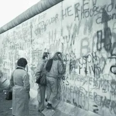 Ludzie oglądają graffiti na Murze Berlińskim, gdy zburzono mur. Widać napisy "Jump over here" i "A house divided cannot stand".