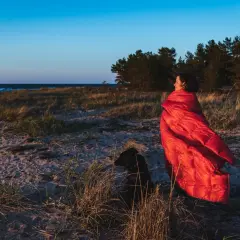 Kobieta w czerwonym śpiworze i pies na plaży. Zastanawiasz się, jaki śpiwór wybrać na nocne wędrówki?