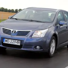 Szary kombi Toyota Avensis T27 1.8 benzyna jedzie po drodze, za nim pola i drzewa.