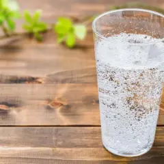 Un verre rempli d'eau gazeuse pétillante, sur une table en bois. Une branche avec des feuilles vertes floues est visible à gauche.