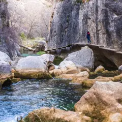 Una persona camina por un puente de madera en la ruta del silencio, rodeada de rocas y un río de aguas cristalinas.