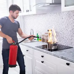 Mann löscht Topfbrand in der Küche mit Feuerlöscher. Dies ist ein Beispiel für Brandklasse F, Fettbrand.