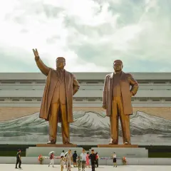 Dwa pomniki przywódców Korei Północnej na tle budynku z malowidłem gór. W tym kraju obowiązują liczne zakazy.