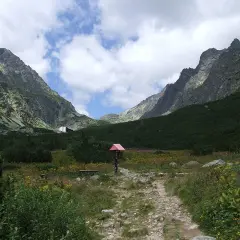 Kamienista ścieżka w górskiej dolinie z zielonymi zboczami i budynkiem w tle.