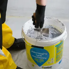 Ręka w czarnej rękawicy miesza łopatką dekoracyjny tynk mozaikowy weber w wiadrze.
