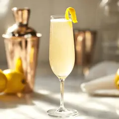 En elegant French 75 med citronskal, bredvid citroner och en kopparskakare.