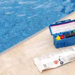Pool test kit and shock treatment next to a blue pool. Learn how to shock a pool for clear, clean water.