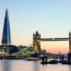 Widok na Tower Bridge i The Shard w Londynie o zmierzchu, z rzeką Tamizą i łodziami w tle.