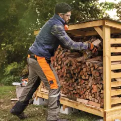Mężczyzna w pomarańczowych rękawicach układa drewno w drewnianej szopie. Obok stoi piła łańcuchowa.