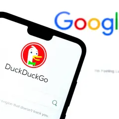 Na ekranie telefonu widoczny jest interfejs wyszukiwarki DuckDuckGo, która jest najlepszą wyszukiwarką internetową dbającą o prywatność. W tle rozmyty napis Google.