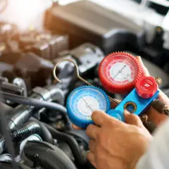 Manometro per ricaricare aria condizionata auto, con due quadranti (rosso e blu) collegati a tubi, in mano a un meccanico davanti al motore.