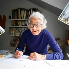 Judith Kerr, lächelnd, zeichnet an ihrem Schreibtisch. Bücherregale im Hintergrund.