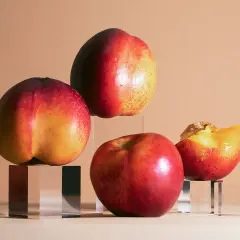 Drei ganze und eine angeschnittene Nektarine auf transparenten Würfeln arrangiert. Die Früchte sind rot-gelb gefärbt und glänzen leicht.