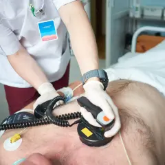 Ratownik medyczny wykonuje kardiowersję pacjentowi leżącemu w łóżku szpitalnym, używając defibrylatora.