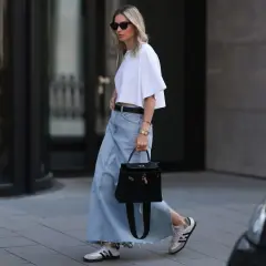 Kobieta w białej bluzce i luźnej, jasnej spódnicy jeansowej plus size, z czarną torebką i białymi sneakersami.