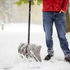 Osoba w czerwonej kurtce i jeansach stoi w śniegu z łopatą do odśnieżania.