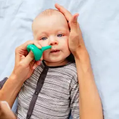 Un bebé con ojos azules recibe un lavado nasal con un aspirador verde. Una mano sujeta su cabeza mientras la otra usa el dispositivo.
