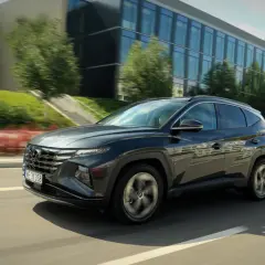 Ciemnoszary Hyundai Tucson 2024 pędzi po drodze, mijając nowoczesny budynek i zielone drzewa.